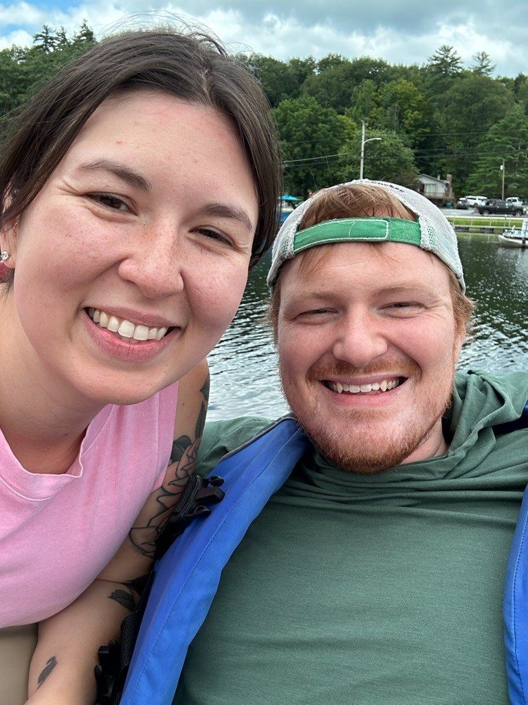 Matt and Chloe smiling in a selfie on a boat, with trees and a lake behind them